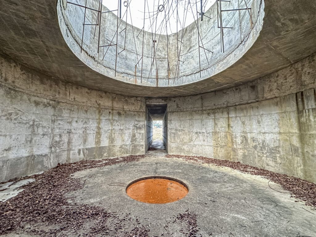 Interior photo of a concrete art installation called The Subrosa Pantheon. It is a circular, concrete room with a large open-air circular opening at the top, a small circular pool in the floor, and a narrow passageway at the back that leads outside.