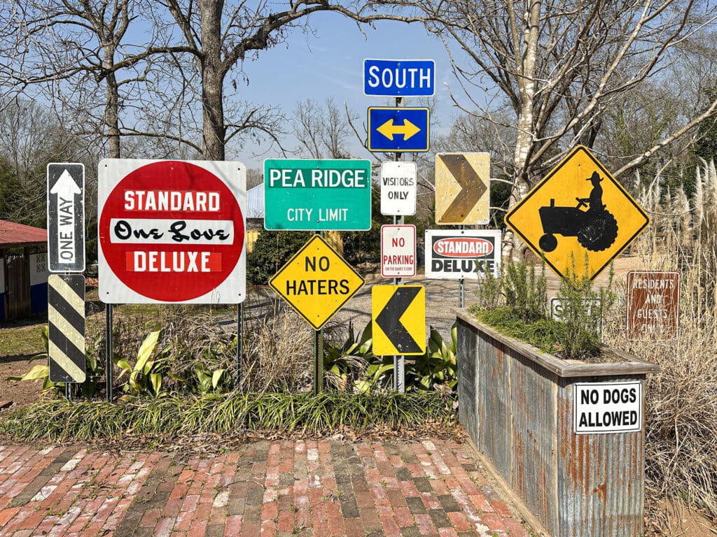 Multiple road signs all collected together and standing in an informal, brick courtyard.