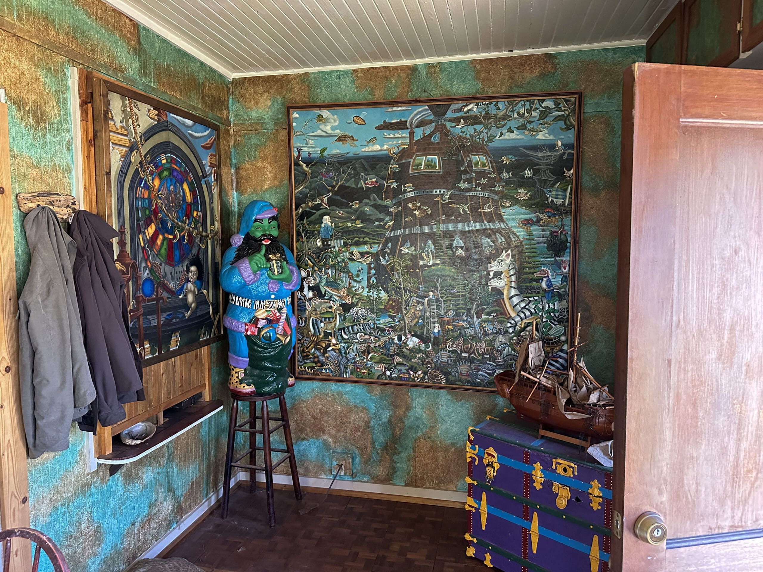 A photo taken inside a home foyer with two jackets hanging at left, two large psychedelic paintings on the walls in the middle, and an outside door open at right.