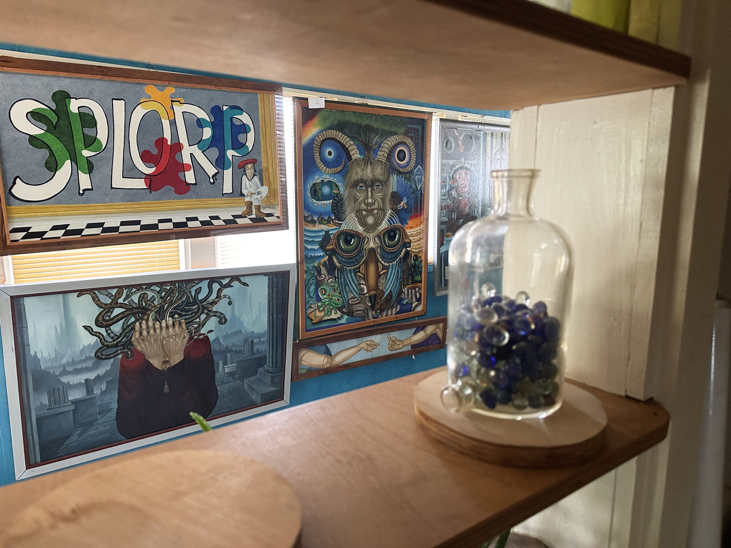 A view between shelves of several paintings: one featuring a crying Medusa, one with a psychedelic hybrid creature, and one with a painter who has left color splotches across giant white letters reading "SPLORP".